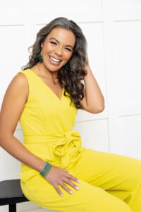 Erin Attere – Teacher A woman in a bright yellow jumpsuit sits smiling on a bench against a white paneled wall, embodying the joyful and welcoming atmosphere of a peaceful retreat at South Shore Resort and Retreats, perfect for a relaxing getaway in West Michigan.| South Shore Resort & Retreats in West Michigan