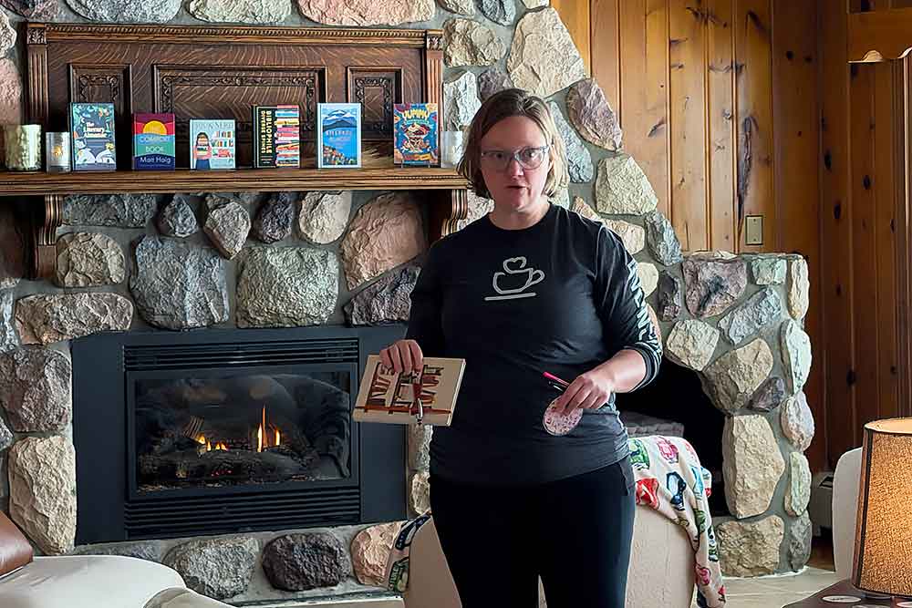 A person stands in a cozy living room with a stone fireplace, showcasing a book. The warm ambiance of the room highlights the charm of a peaceful retreat, ideal for a relaxing getaway at South Shore Resort and Retreats in West Michigan.| South Shore Resort & Retreats in West Michigan