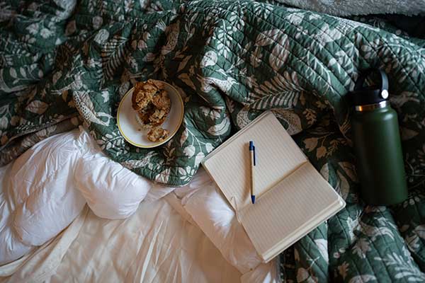 A cozy bed with a green leafy quilt, a notebook with a pen, a plate of cookies, and a water bottle evoke a peaceful retreat at South Shore Resort and Retreats, perfect for a relaxing getaway in West Michigan.| South Shore Resort & Retreats in West Michigan