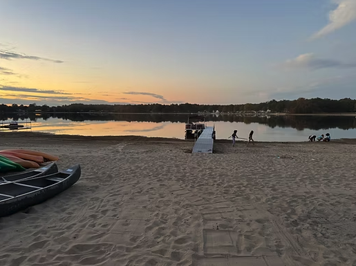 A tranquil beach at sunset with a dock extending into calm water, kayaks on the sand, and people enjoying the serene view, perfect for a relaxing getaway at South Shore Resort and Retreats, a beautiful lakefront retreat in West Michigan.| South Shore Resort & Retreats in West Michigan