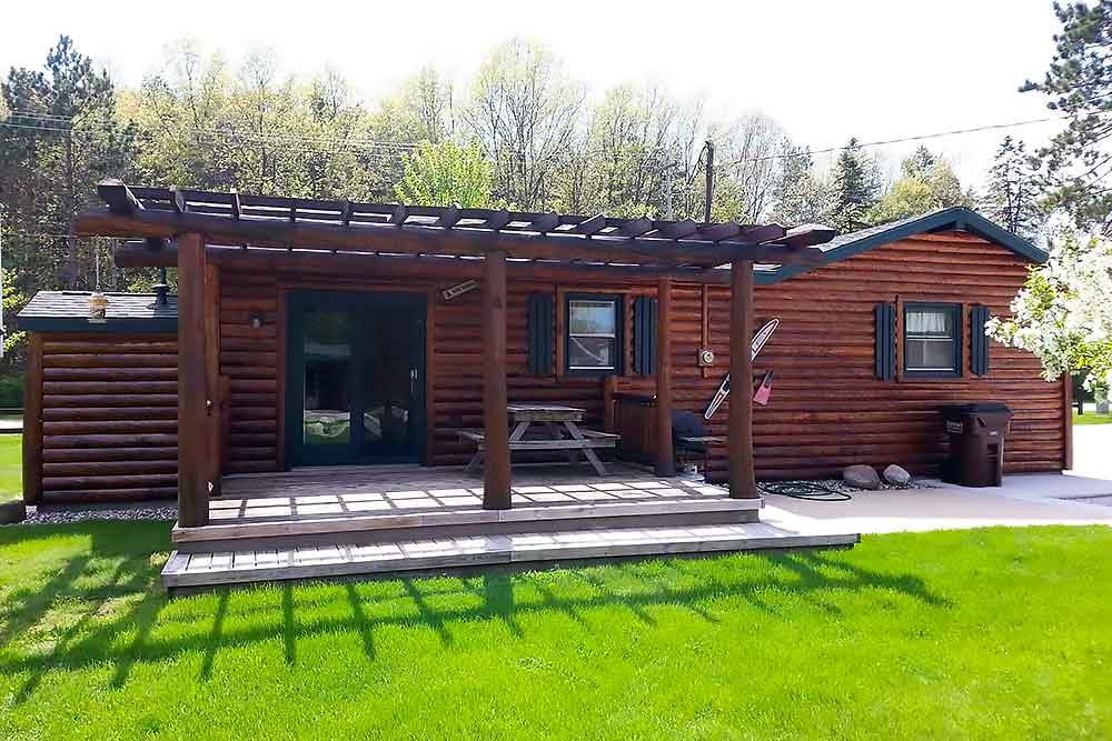 A cozy log cabin with a pergola stands against a lush green backdrop, ideal for a family vacation at a peaceful retreat. Enjoy serene moments at South Shore Resort and Retreats in West Michigan, perfect for a relaxing getaway near Lake Michigan.| South Shore Resort & Retreats in West Michigan