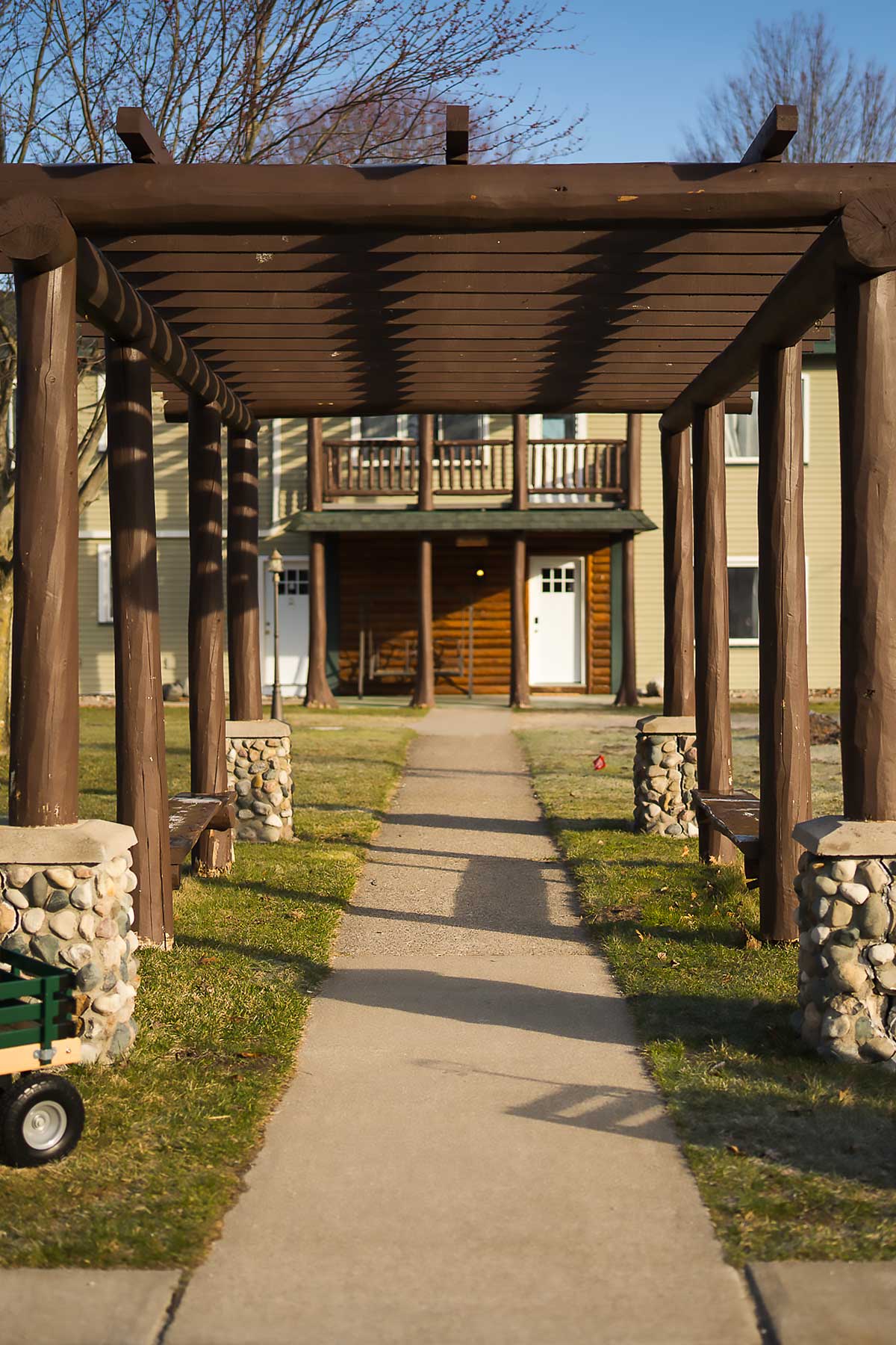 A wooden pergola with stone pillars lines a sunny path leading to a cozy lodge, ideal for a relaxing getaway at South Shore Resort and Retreats in West Michigan, offering peaceful and scenic lodging.| South Shore Resort & Retreats in West Michigan