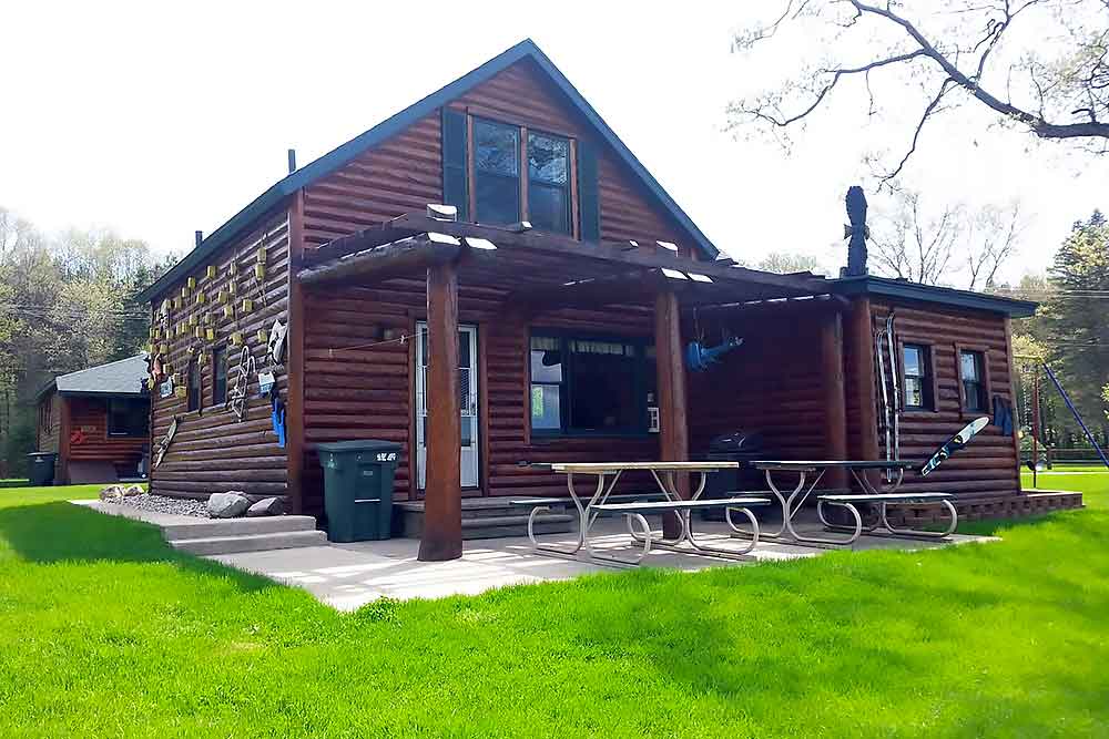 A cozy log cabin with a wooden pergola and picnic tables sits in lush green surroundings, ideal for a peaceful retreat at South Shore Resort and Retreats, offering a serene West Michigan vacation rental experience.| South Shore Resort & Retreats in West Michigan