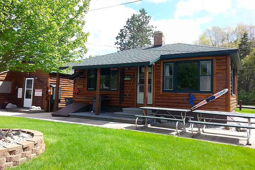 A cozy log cabin with a picnic table sits under a bright sky, surrounded by lush greenery, offering a perfect setting for a family vacation at South Shore Resort and Retreats, a premier West Michigan vacation rental destination.| South Shore Resort & Retreats in West Michigan