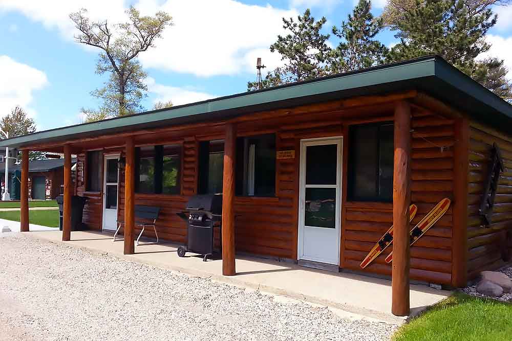 A cozy log cabin with a green roof at South Shore Resort and Retreats, featuring a grill and Adirondack chairs on the porch, set against a backdrop of lush trees and blue sky, perfect for a relaxing getaway in West Michigan.| South Shore Resort & Retreats in West Michigan