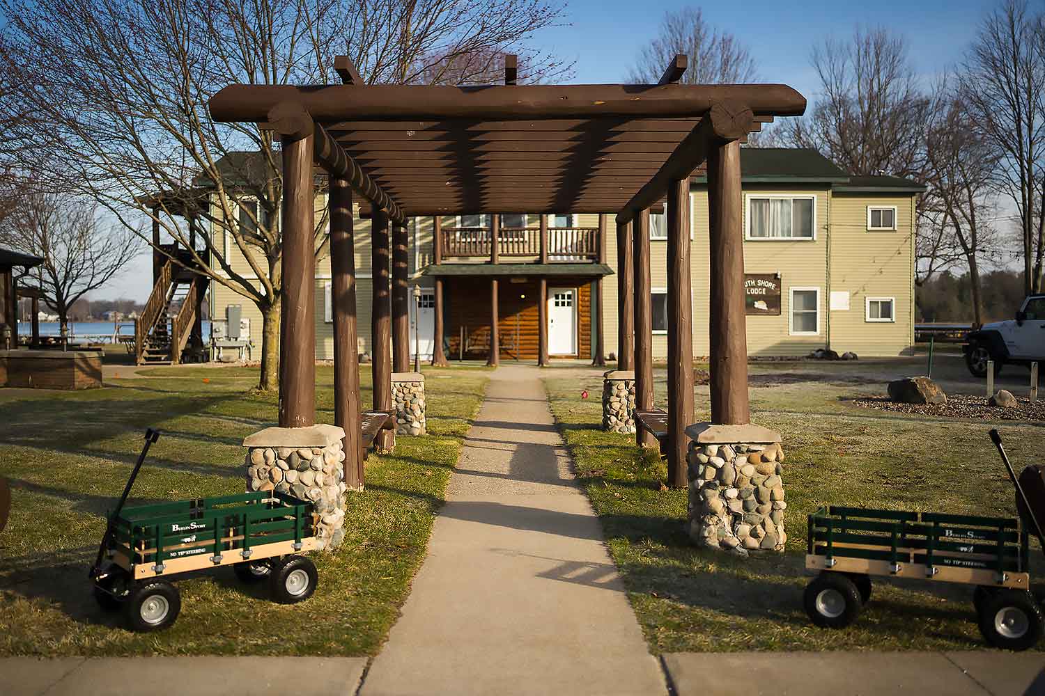 A wooden pergola leads to a cozy lodge at South Shore Resort and Retreats, surrounded by trees and carts on a sunny day, offering a peaceful retreat for families and groups seeking a relaxing getaway in West Michigan.| South Shore Resort & Retreats in West Michigan