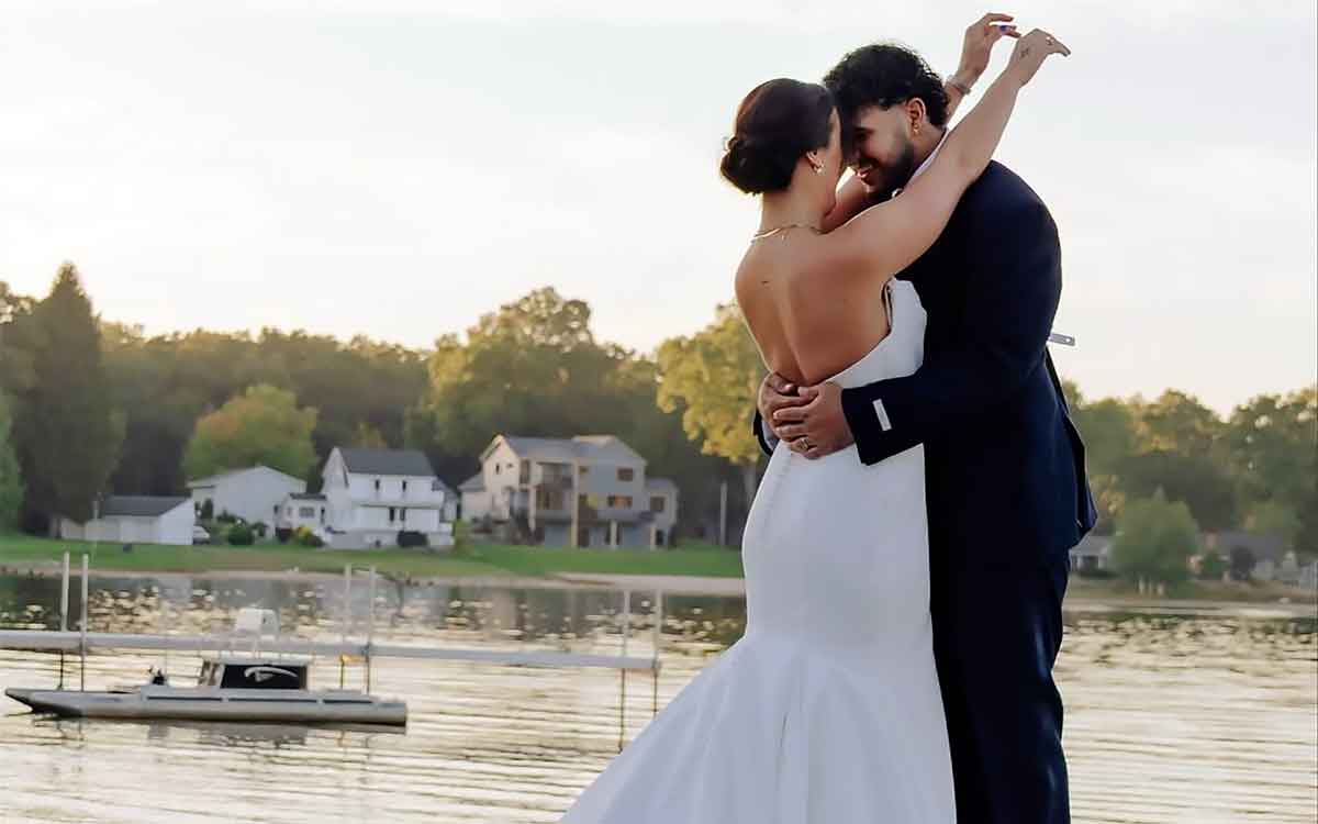 A couple in wedding attire shares a romantic embrace by a serene lake at sunset, with lush greenery and cozy homes in the background, capturing the essence of a peaceful retreat at South Shore Resort and Retreats.| South Shore Resort & Retreats in West Michigan